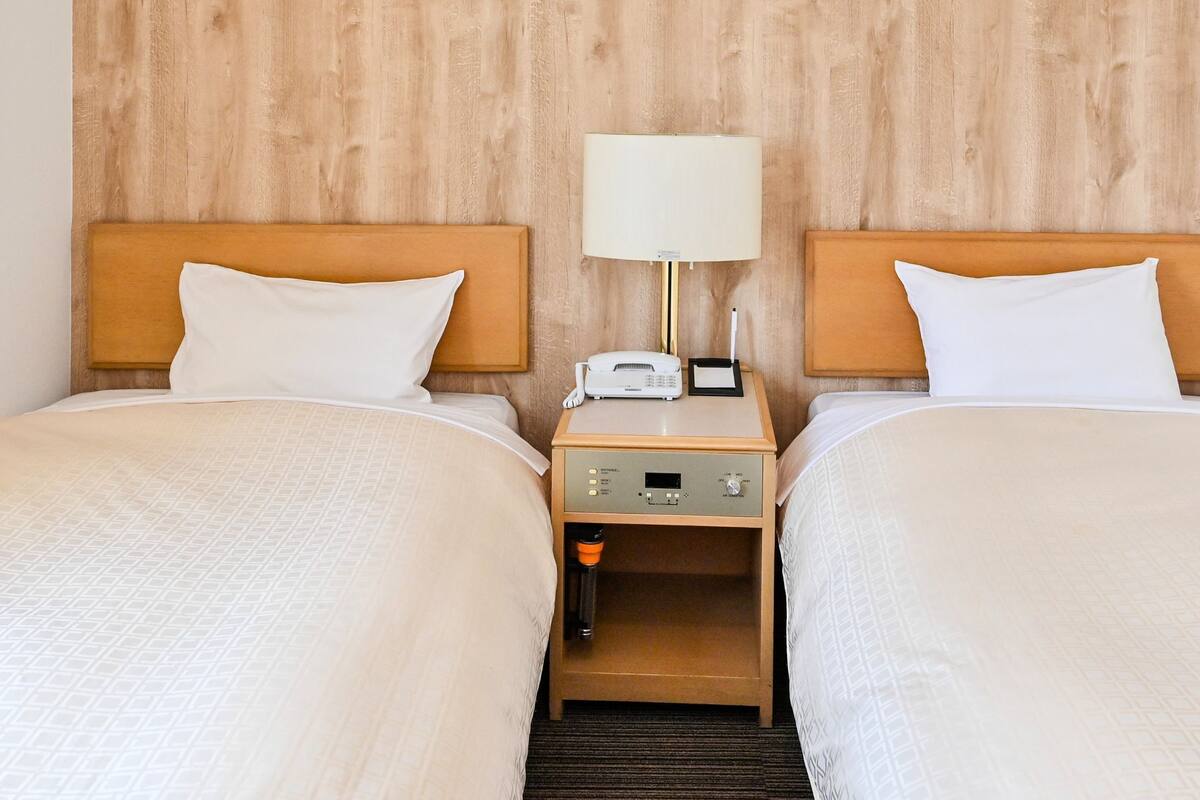A hotel room with two beds, wooden headboards, white bedding, a bedside table with a lamp, a phone, and a thermostat.