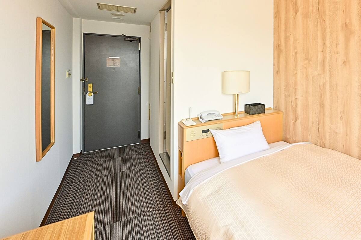 A hotel room with a bed, a bedside table with a lamp, a telephone, and a door.