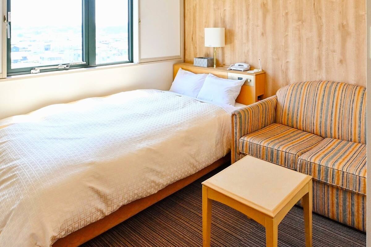 A hotel room with a bed, a striped sofa, a small table, a nightstand with a lamp, and a window with a view.