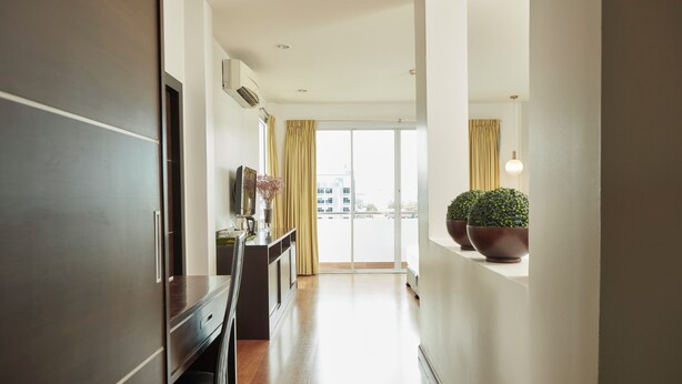 A modern hotel corridor with a wooden floor, a kitchenette, a large window with curtains, and potted plants. - Rhienchai Place (Surat Thani)