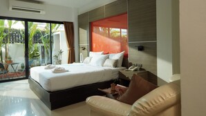 A modern bedroom with a large bed, a bedside table with a phone, and a view of a balcony with plants. - Rhienchai Place (Surat Thani)