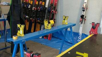 A ski equipment storage area with skis and ski poles on racks, a blue workbench, and red and yellow clamps on the floor.