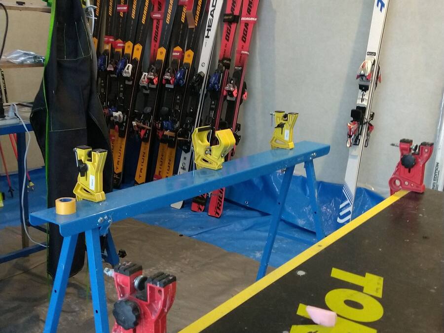 A ski equipment storage area with skis and ski poles on racks, a blue workbench, and red and yellow clamps on the floor.
