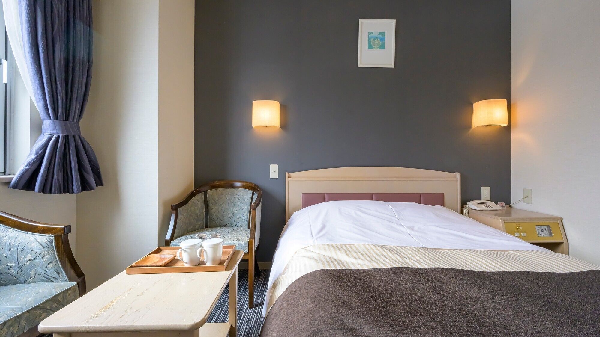 A hotel room with a bed, bedside table with a tray of cups, a chair, a nightstand with a phone, and two wall-mounted lamps.