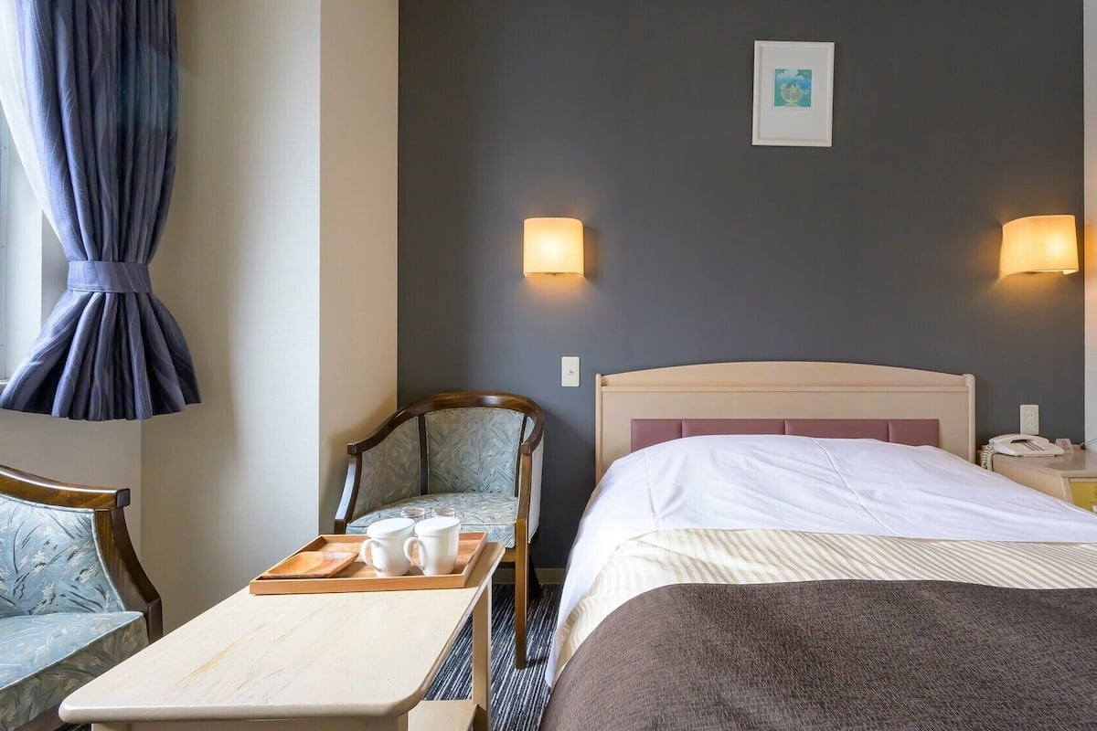 A hotel room with a bed, bedside table with a tray of cups, a chair, a nightstand with a phone, and two wall-mounted lamps.