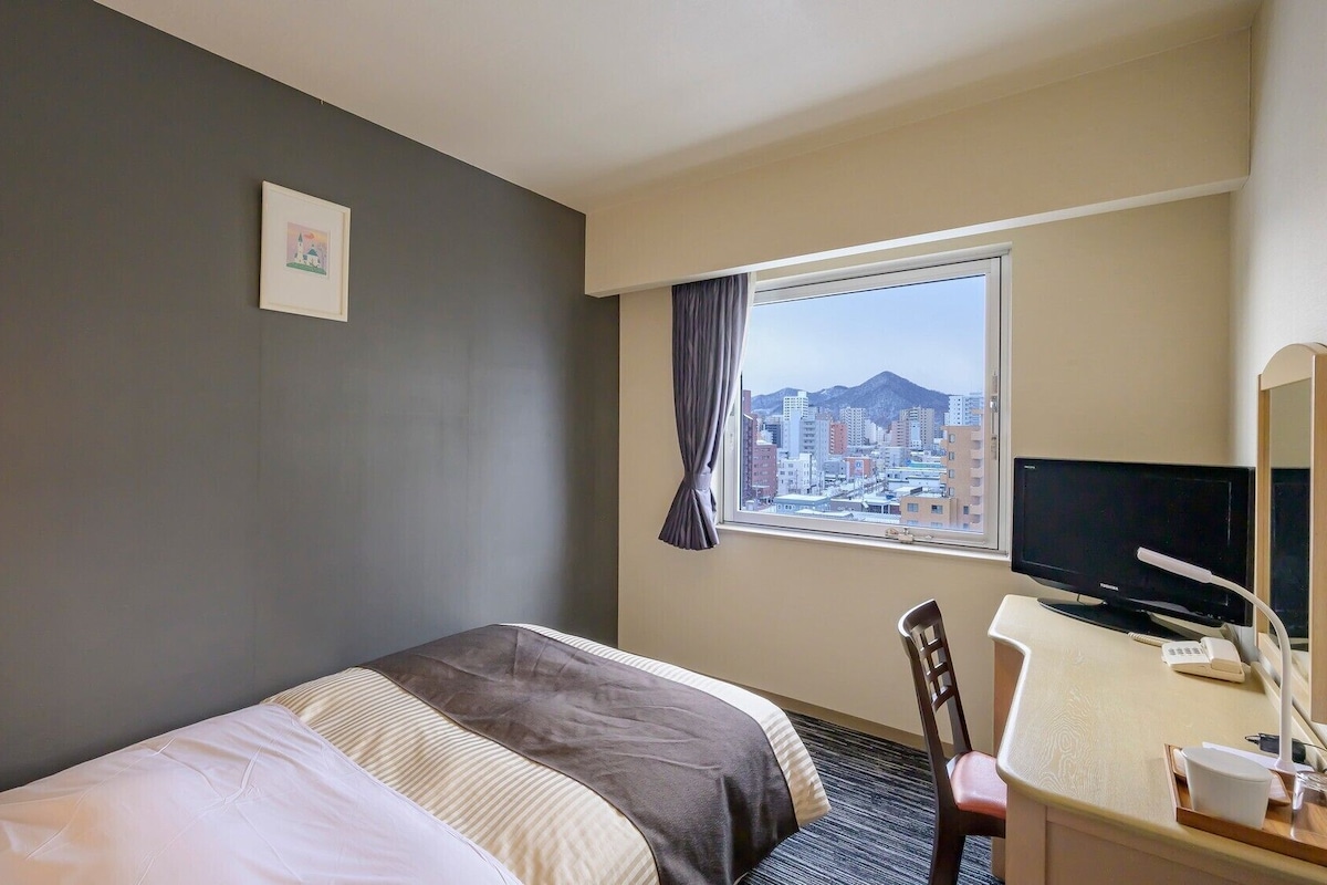 A hotel room with a bed, a desk with a computer, a chair, a window with a city view, and a wall-mounted TV.