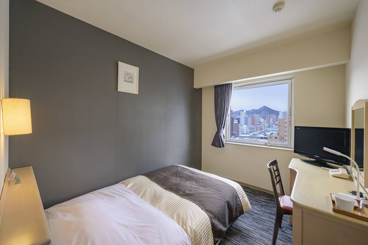 A hotel room with a bed, a desk with a computer, a chair, a lamp, and a window with a city view.