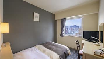 Habitación de hotel con cama, escritorio con computadora, silla, lámpara y ventana con vista a la ciudad.