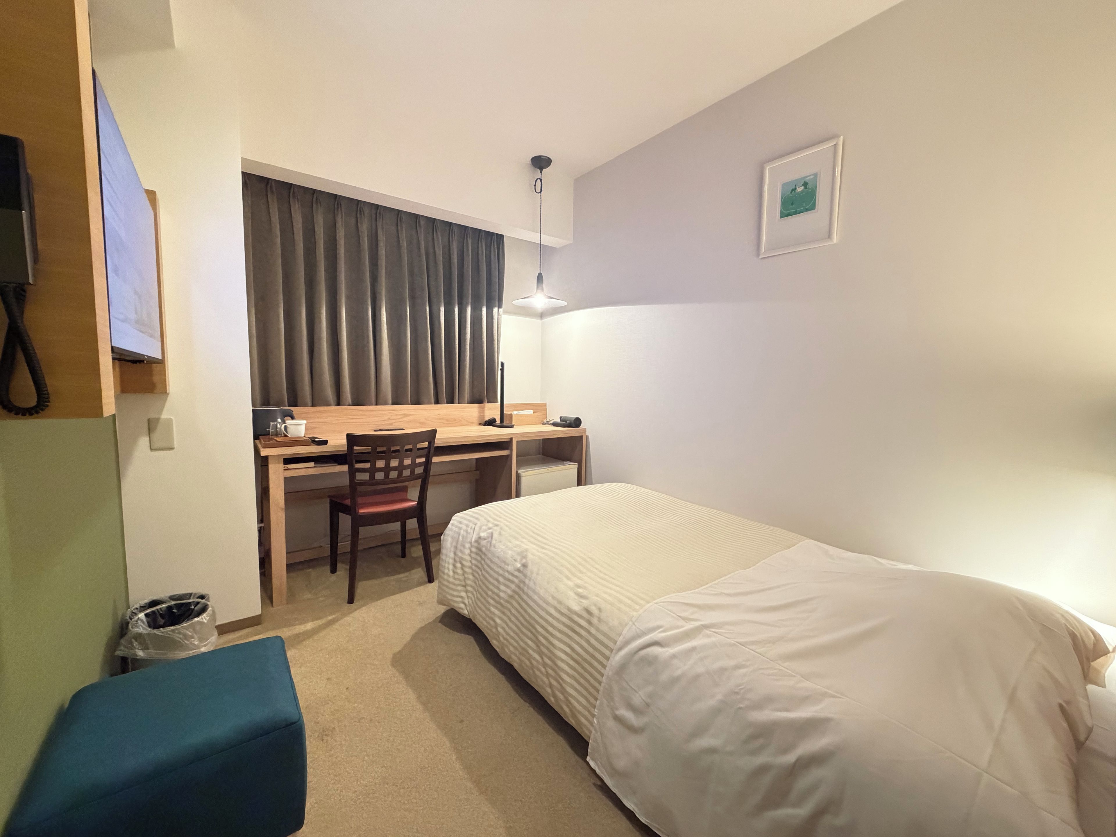A hotel room with a single bed, a desk, a chair, a small table, a trash bin, and a wall-mounted telephone.