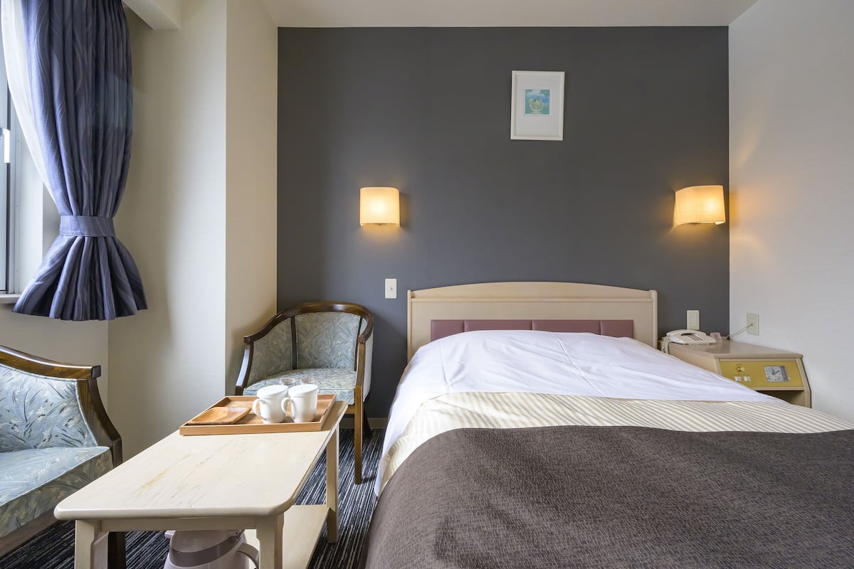 A hotel room with a bed, a chair, a small table with a tray, and two wall-mounted lamps.