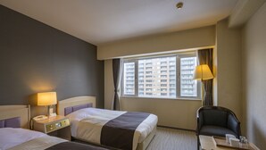 Quarto de hotel com duas camas, uma mesa, uma cadeira, um abajur e janela com vista para edifícios.