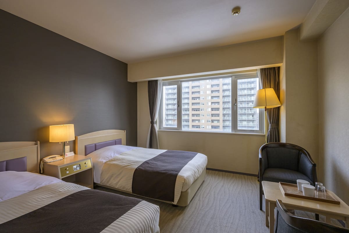 A hotel room with two beds, a desk, a chair, a lamp, and a window with a view of buildings.
