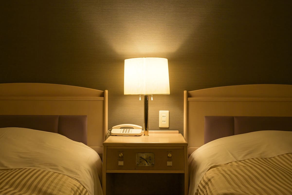 A hotel room with two beds, a nightstand with a lamp, a phone, and a clock.