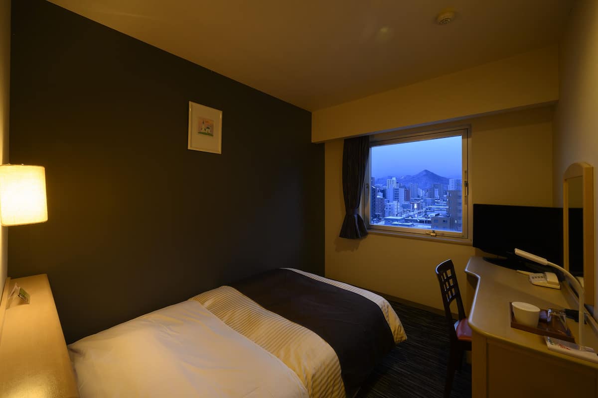 A hotel room with a bed, a desk, a chair, a lamp, a window with a city view, and a small picture on the wall.