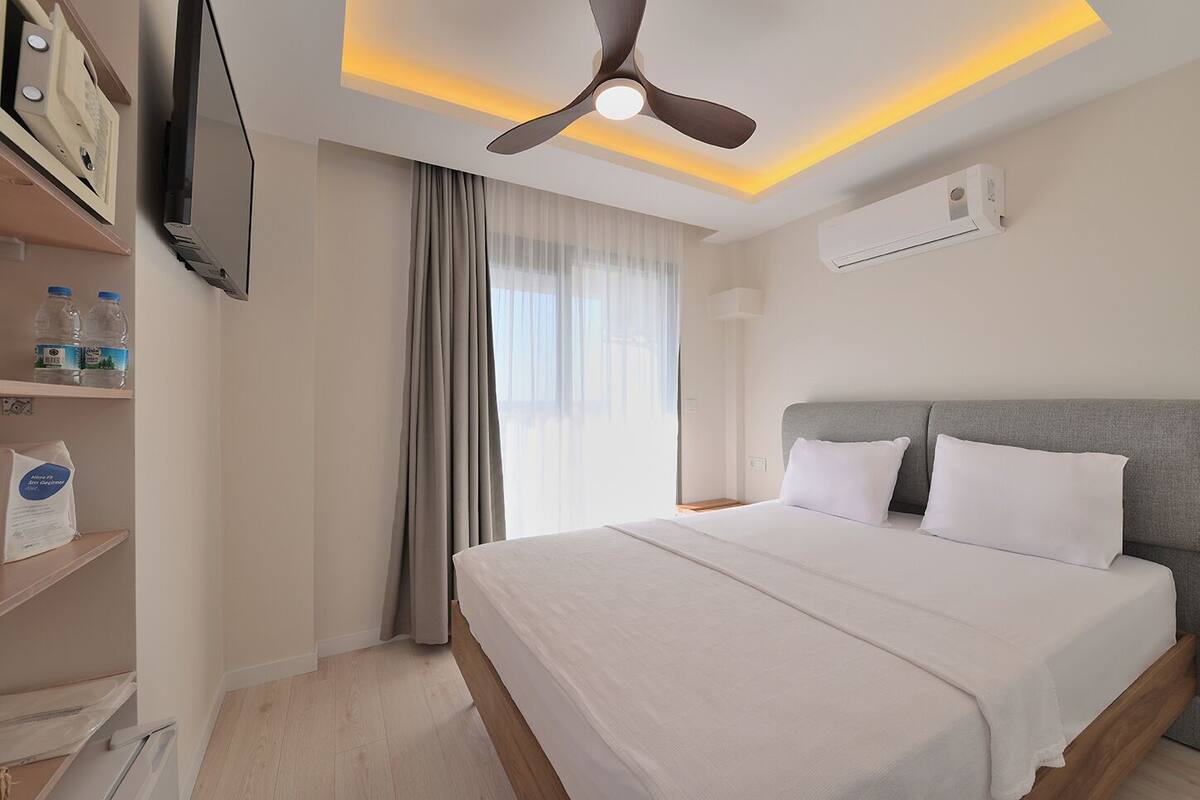A hotel room with a bed, a ceiling fan, an air conditioning unit, a TV, and a window with curtains.