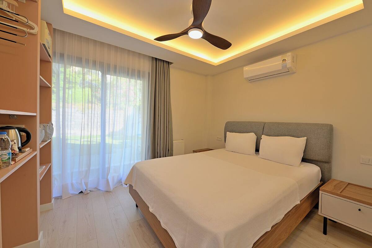 A hotel room with a large bed, a ceiling fan, an air conditioning unit, a bedside table, and a wardrobe.