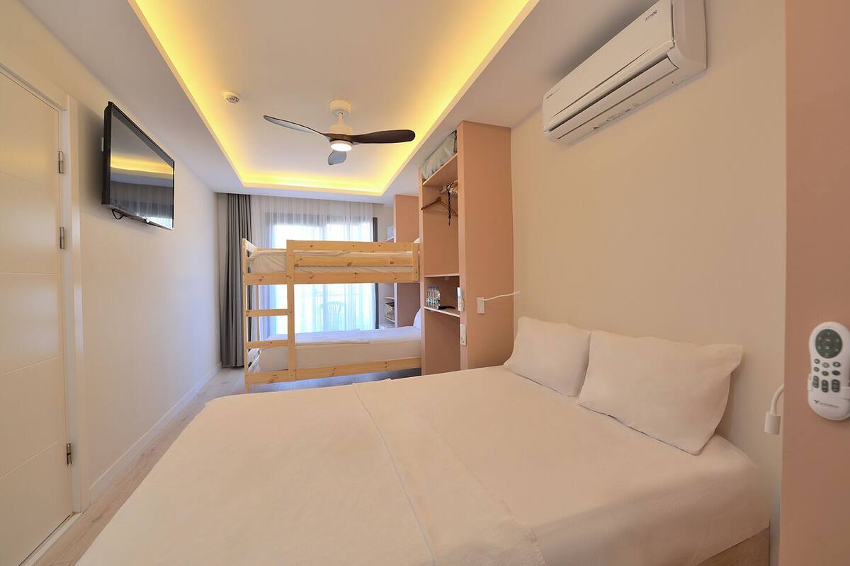 A hotel room with a bunk bed, a ceiling fan, an air conditioning unit, a TV mounted on the wall, and a bedside table with a remote control.