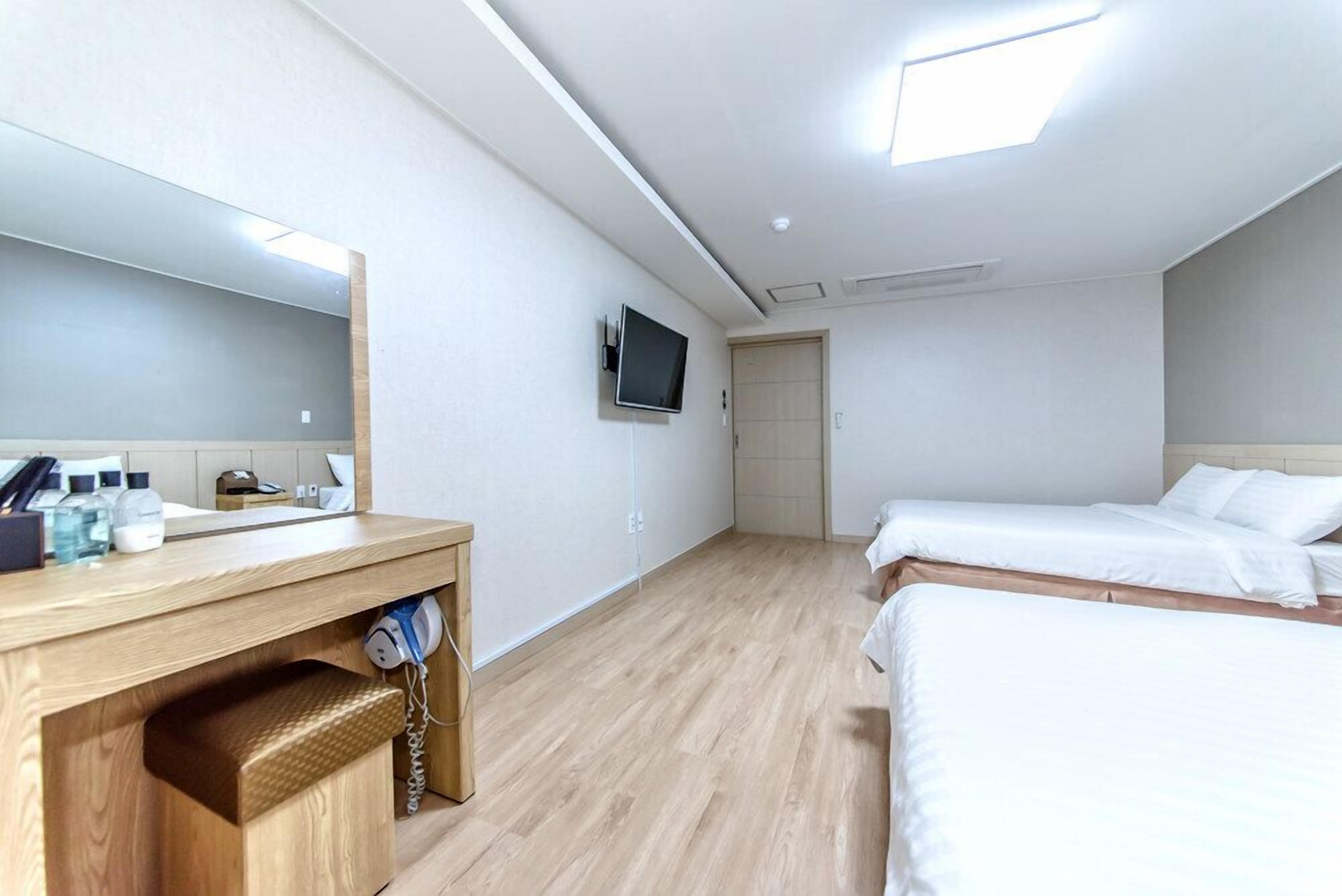 A hotel room with a wooden desk, a large mirror, a TV mounted on the wall, two beds, and a door leading to another room.