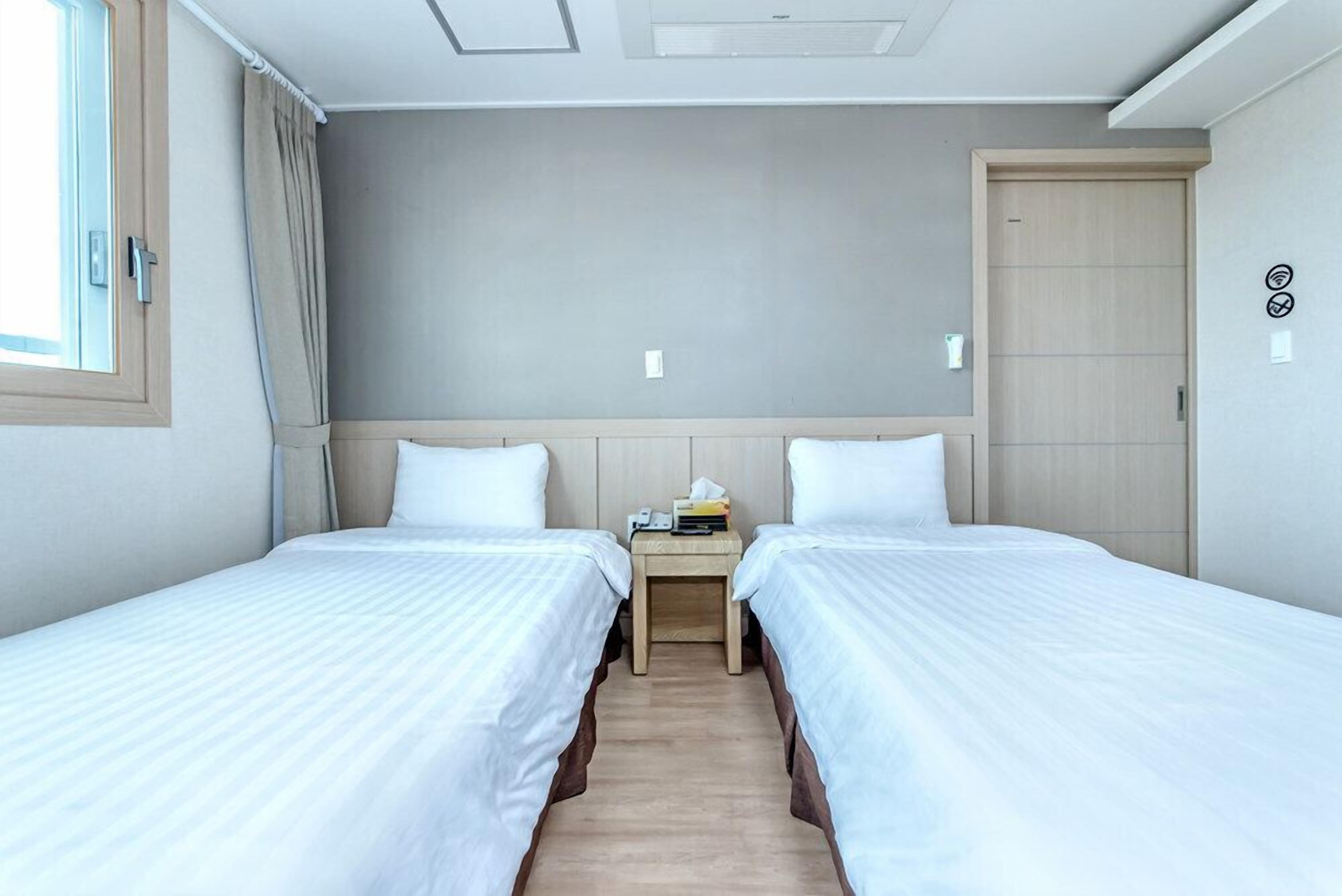 A hotel room with two single beds, a wooden nightstand with a phone and tissues, a window with curtains, and a door with a light switch.