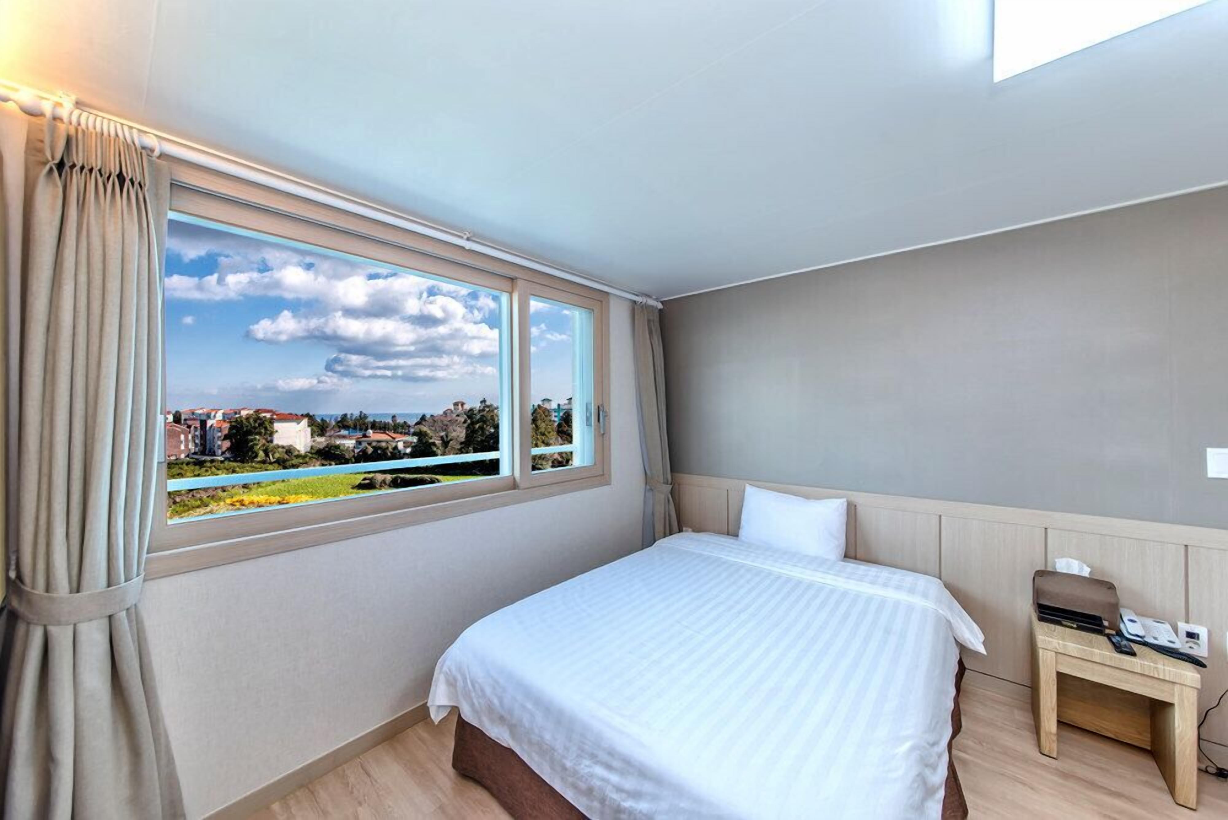 Ein Hotelzimmer mit großem Fenster, einem Bett, einem Nachttisch und Blick auf die Stadt.
