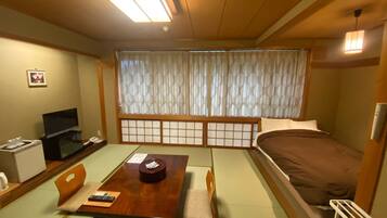 Una habitación tradicional japonesa con suelo de tatami, una mesa baja con juego de té, una cama con futón y un televisor sobre un mueble bajo.