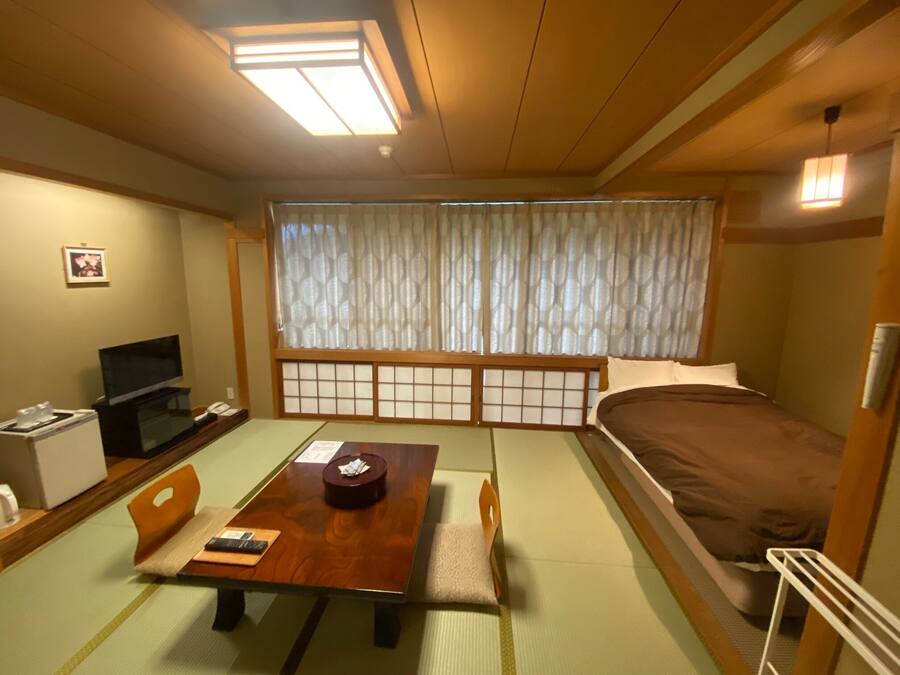 Una habitación tradicional japonesa con suelo de tatami, una mesa baja con juego de té, una cama con futón y un televisor sobre un mueble bajo.