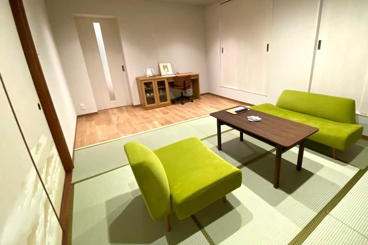 A room with tatami flooring, a wooden table, two green chairs, and a sofa.