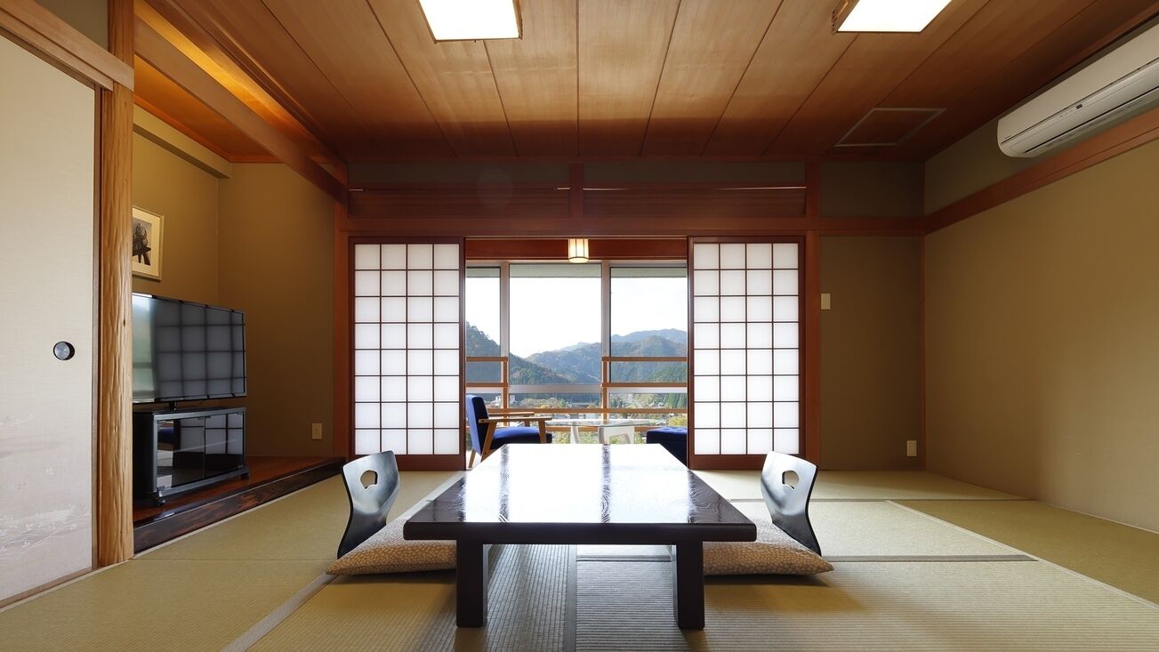 A tatami mat room with a low table, chairs, a television, and a view of mountains through large windows.