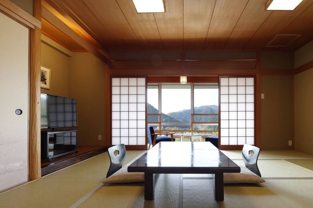 A tatami mat room with a low table, chairs, a television, and a view of mountains through large windows.