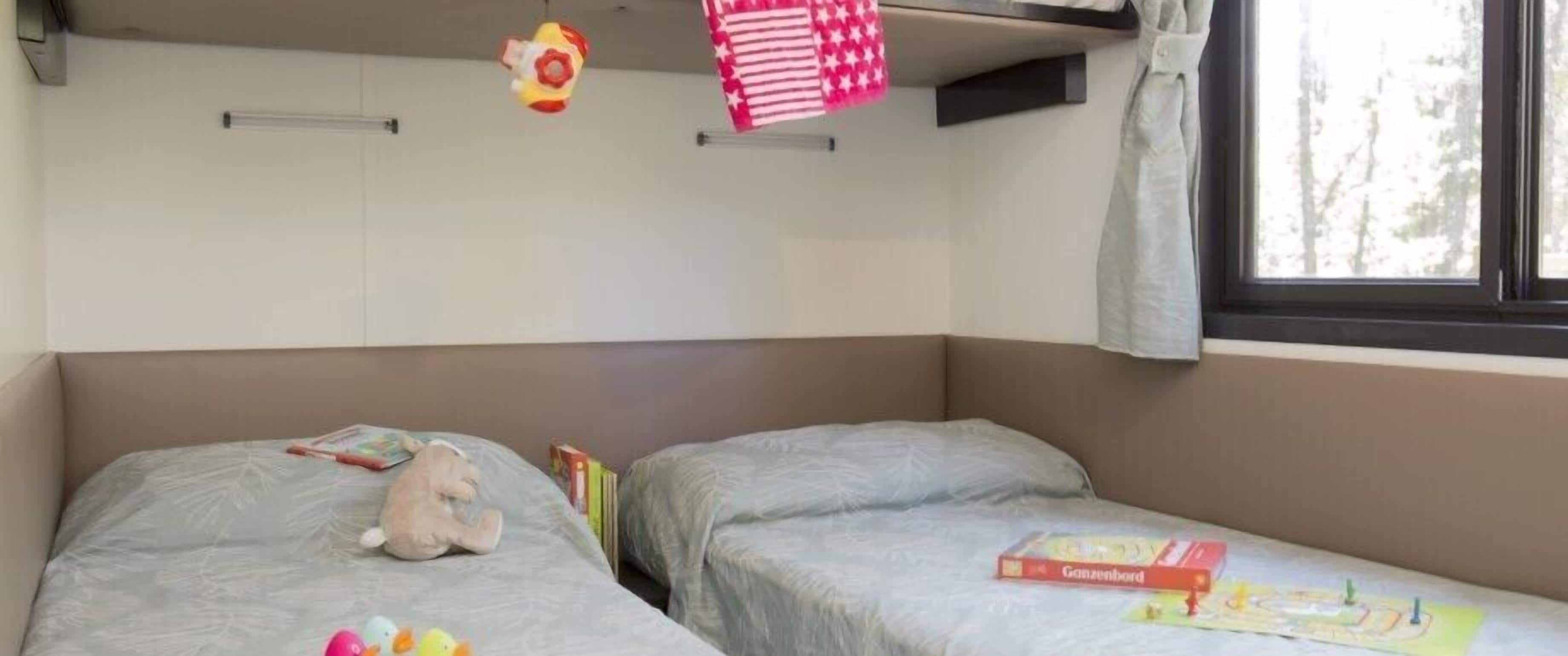 Une chambre avec deux lits, chacun recouvert d’une couverture et doté d’oreillers gris. On y trouve des jouets et des livres sur les lits, ainsi qu’une fenêtre donnant sur des arbres.