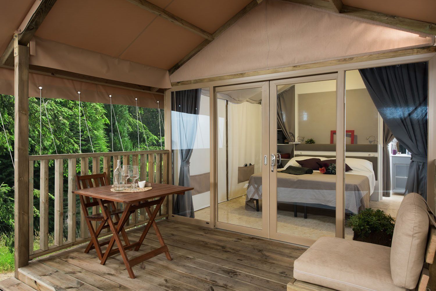 A wooden deck with a table and chairs, a sliding glass door leading to a bedroom with a bed and a bedside table.