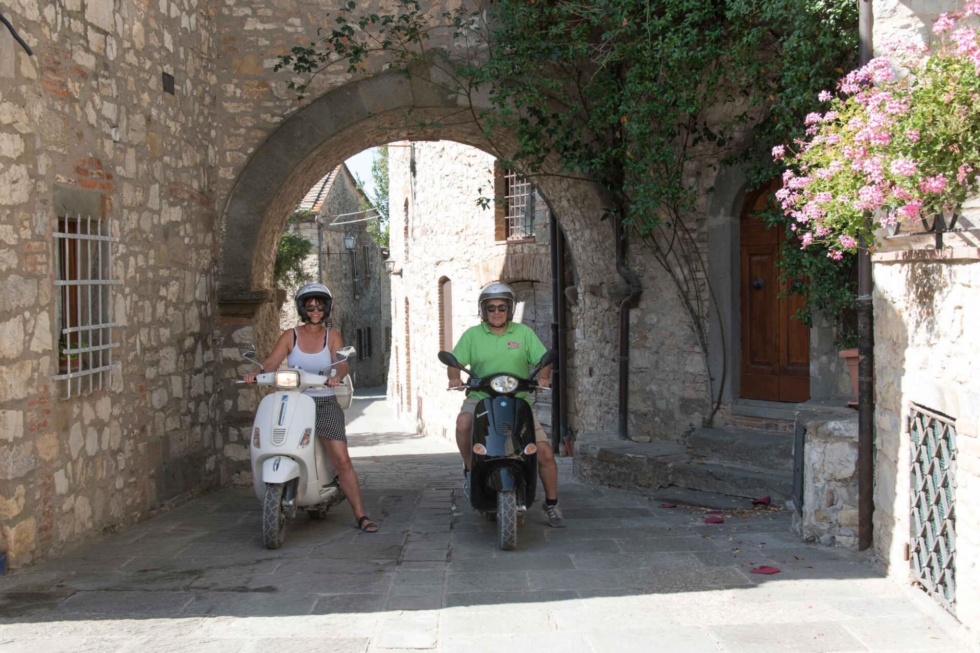 Deux personnes sur des scooters dans une rue étroite bordée de bâtiments en pierre et surmontée d’une arche.