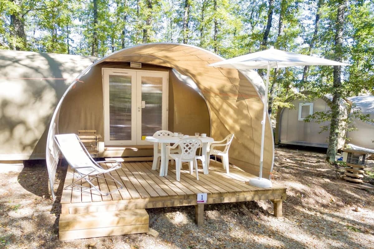 Een gezellige tent buiten met een houten terras, een tafel en stoelen, omringd door bomen.