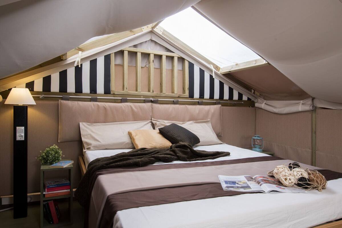Een tent met een bed, een lamp, een klein tafeltje met een plantje en een tijdschrift op het bed.