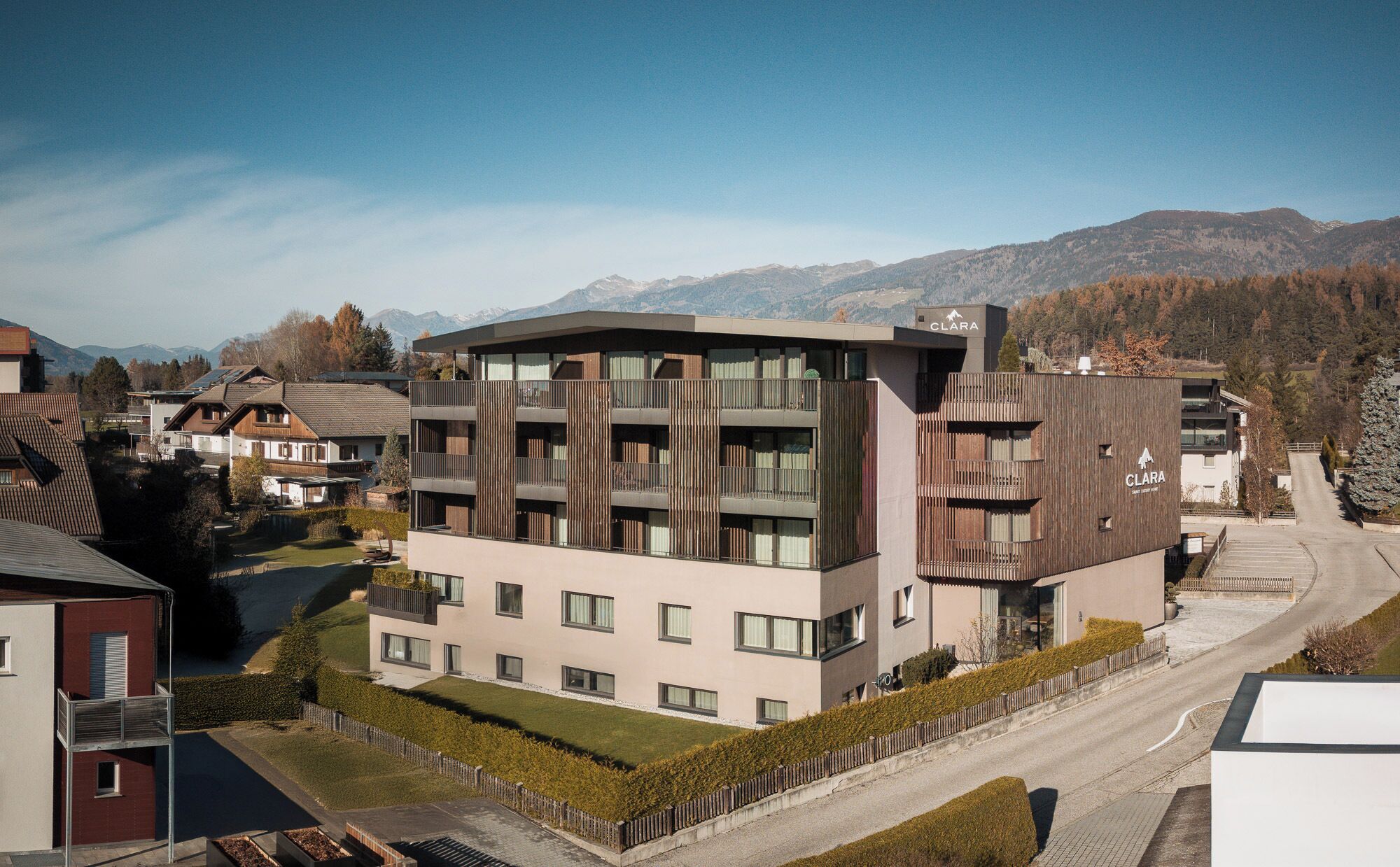 A modern multi-story building with balconies, surrounded by a residential area and mountains in the background.