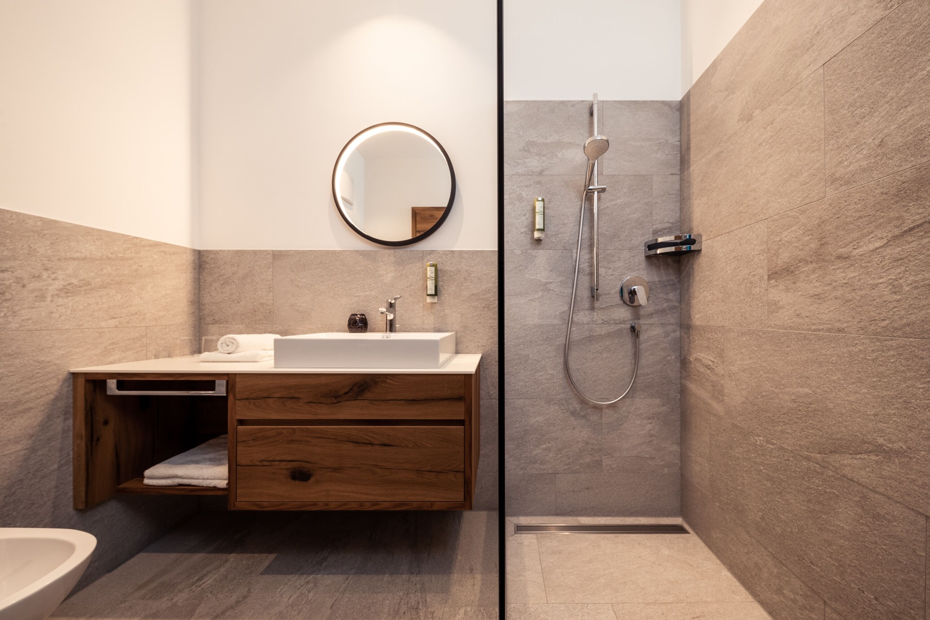 A modern bathroom with a wooden vanity, a round mirror, and a walk-in shower.