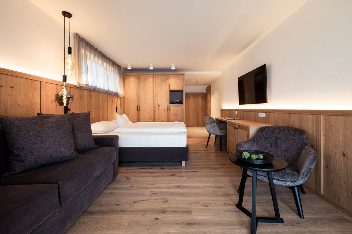 Een moderne, goed verlichte kamer met een bed, een bank, een eettafel met stoelen en een aan de muur bevestigde televisie.
