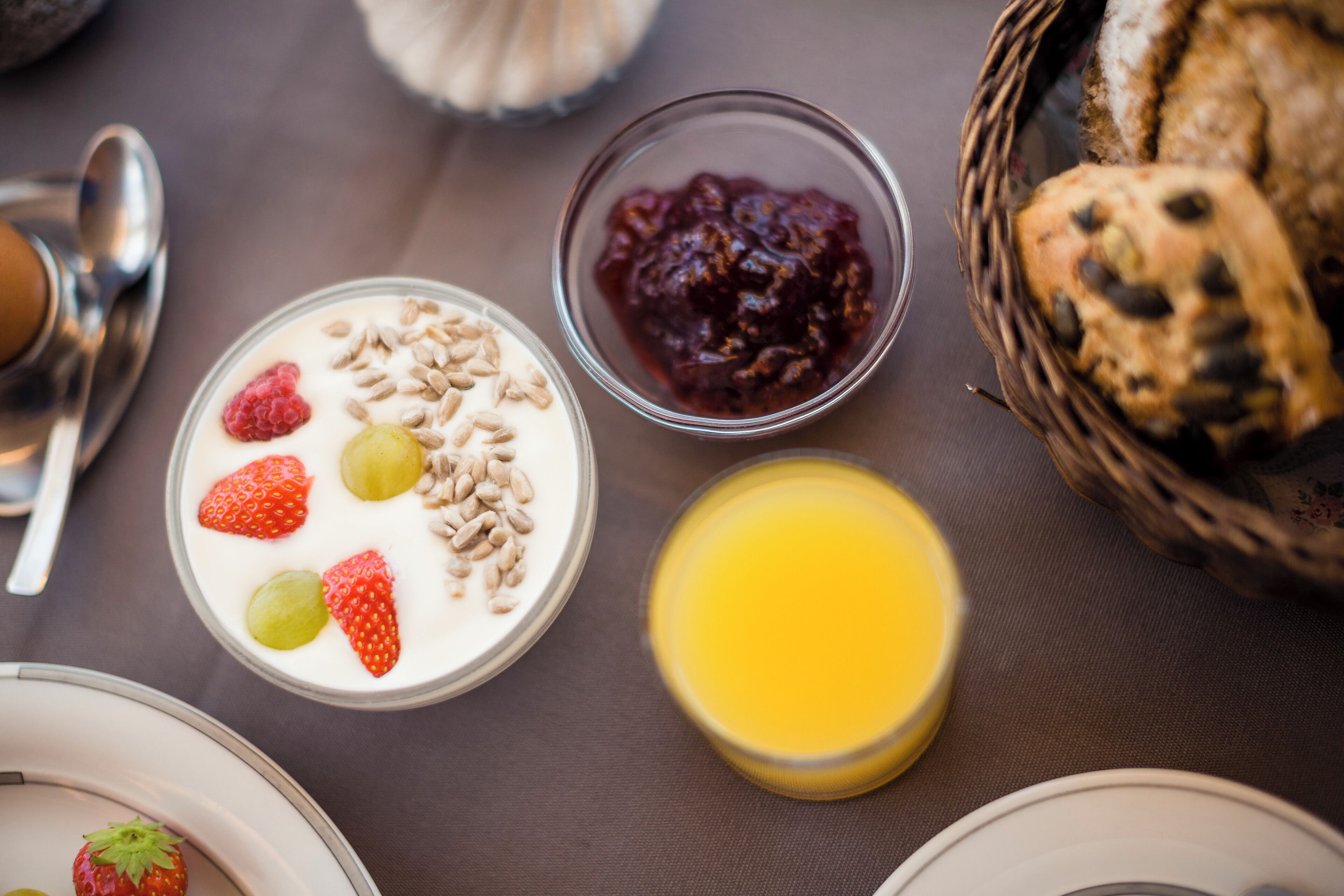 A breakfast setting with a bowl of yogurt topped with strawberries and grapes, a glass of orange juice, and a basket of bread.