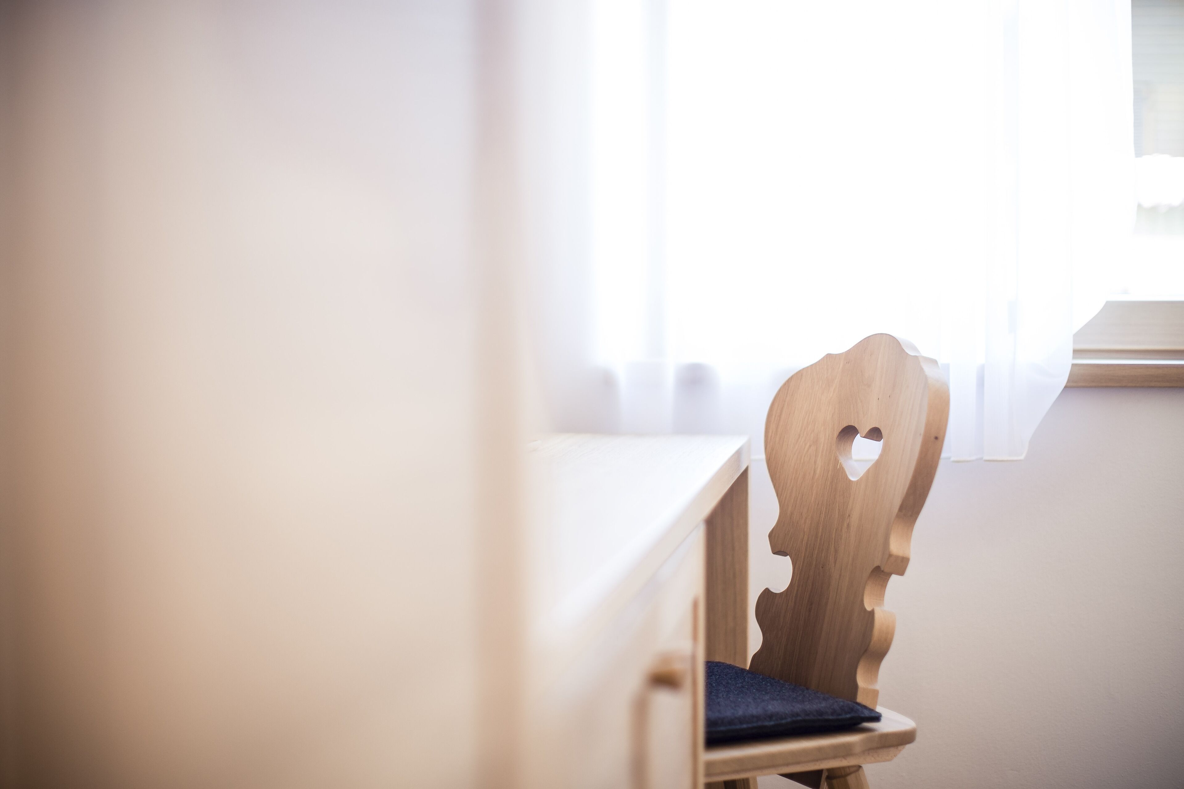 Une chaise en bois avec une découpe en forme de cœur dans le dossier, placée à côté d’un bureau blanc dans une pièce dotée d’une fenêtre.