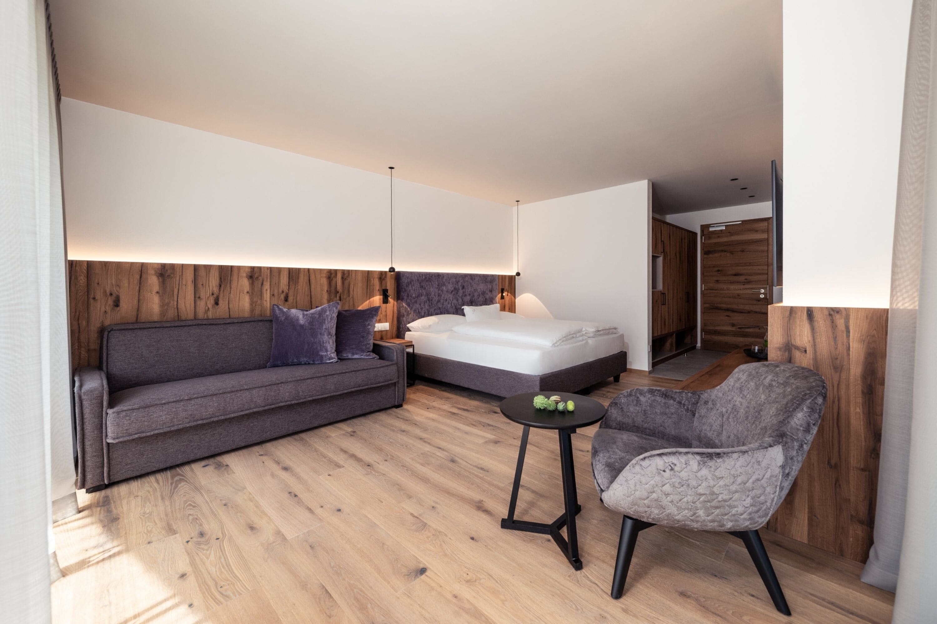 A modern hotel room with a wooden floor, a grey sofa, a bed, a small round table with a lamp, and a grey armchair.