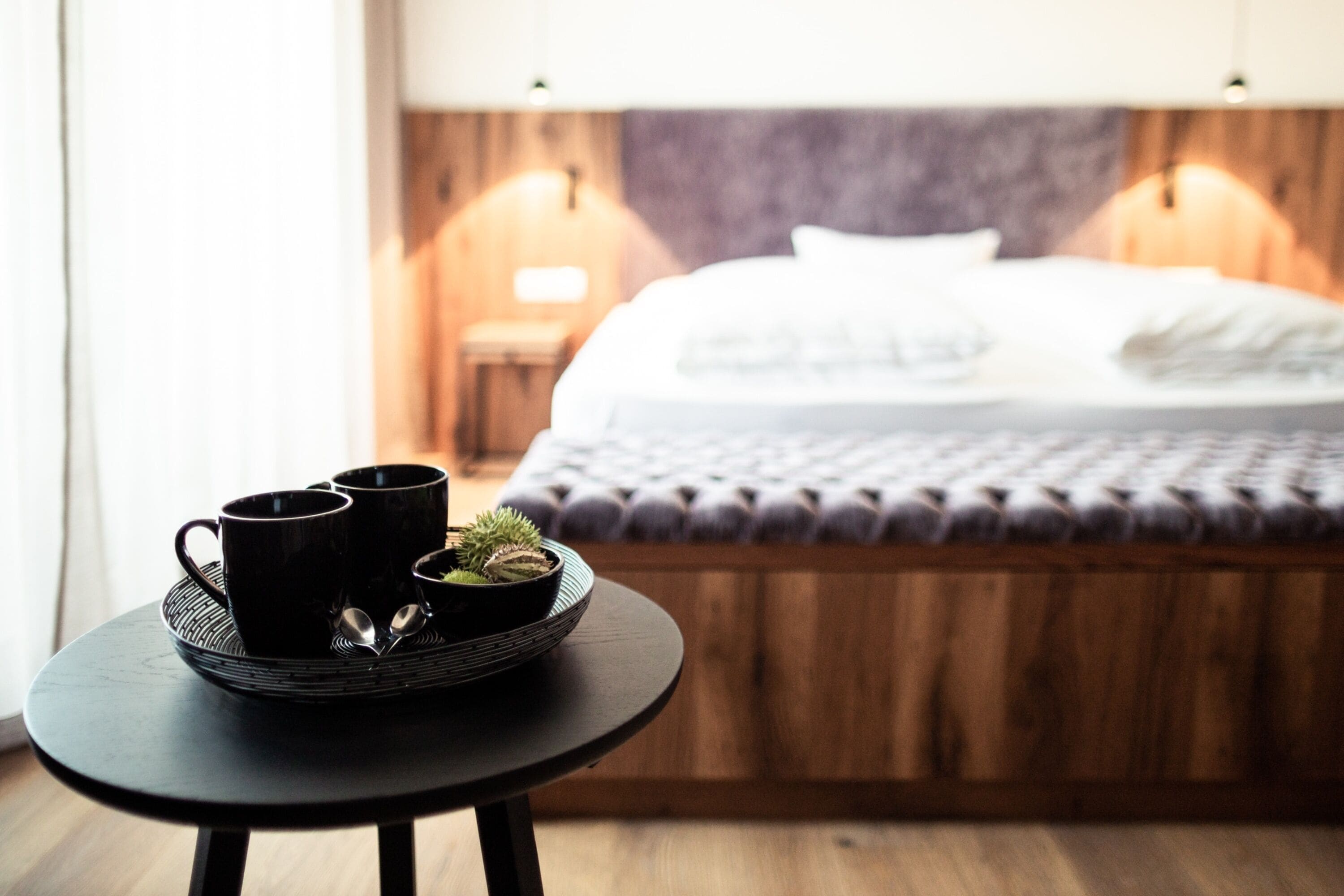 Una tavola da colazione ordinata con tazze nere e una piccola pianta, un letto con testiera in legno e un comodino con una lampada in una camera d'albergo moderna.