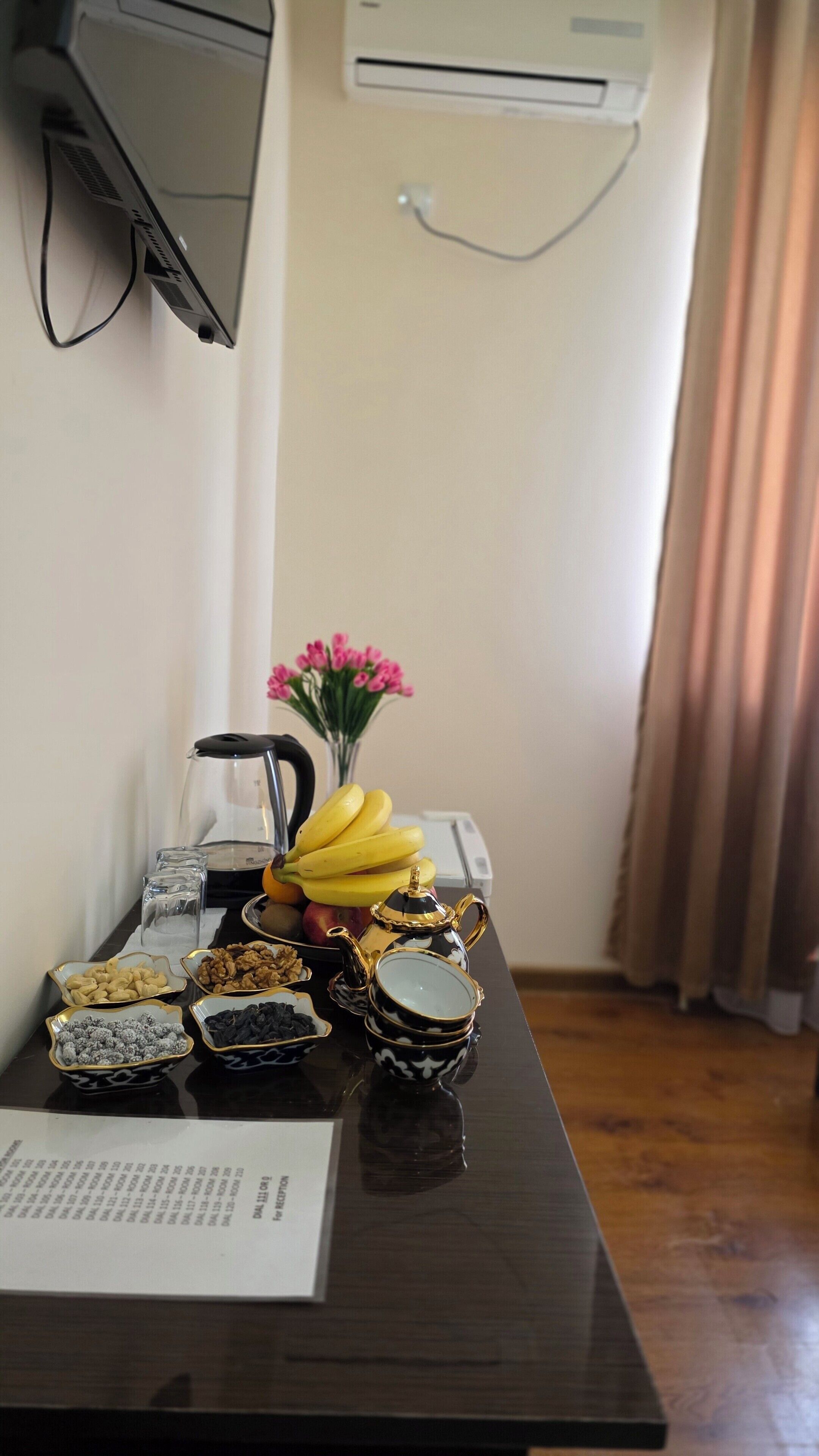 A hotel room with a desk, a flower vase, a kettle, and a fruit bowl.