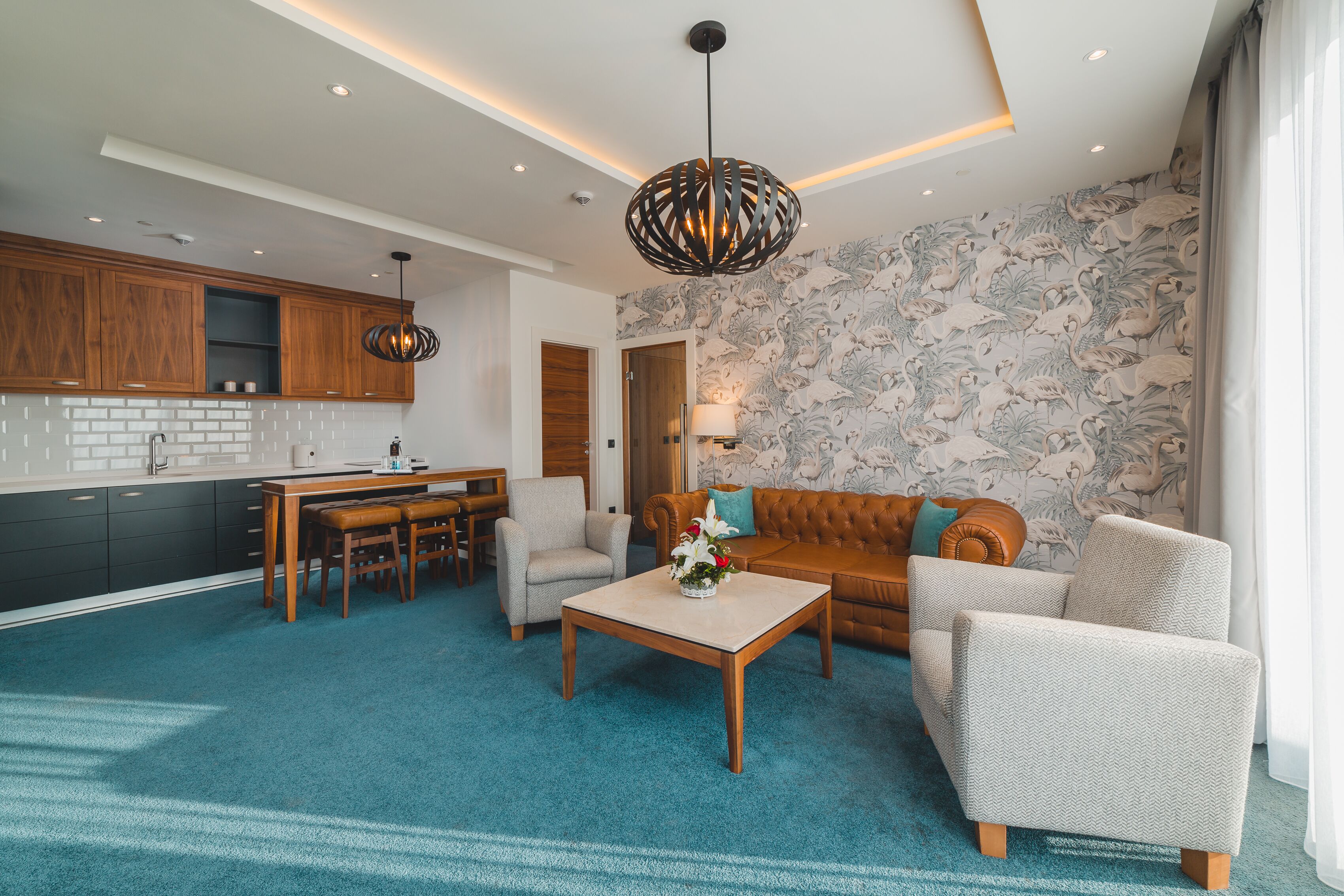 A modern living room with a sofa, armchair, and coffee table. There is a kitchen area with wooden cabinets and a dining table.