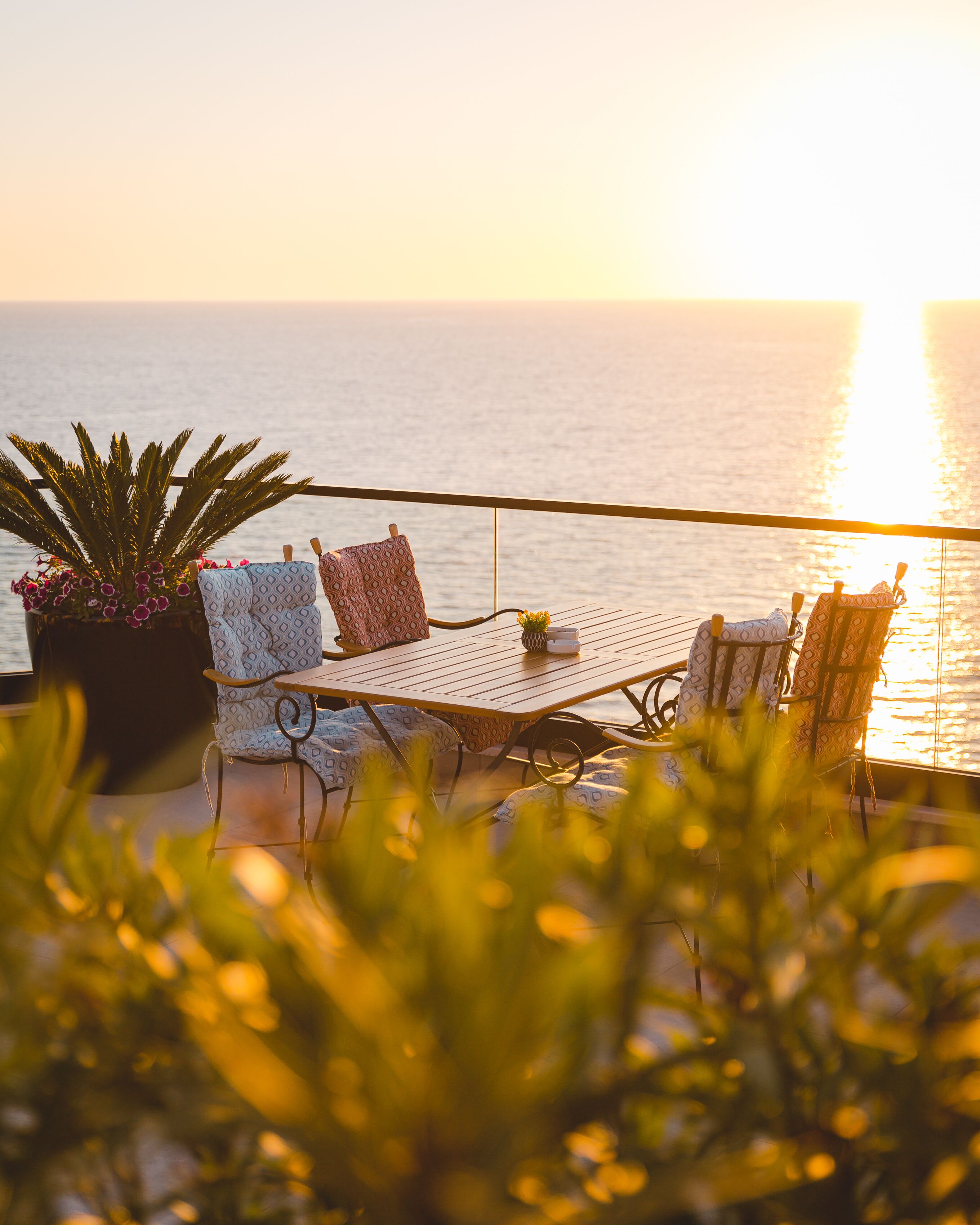 A terrace with a table and chairs, a potted plant, and a view of the sea at sunset.
