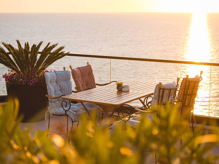 Um terraço com mesa e cadeiras, um vaso de planta e vista para o mar ao pôr do sol.