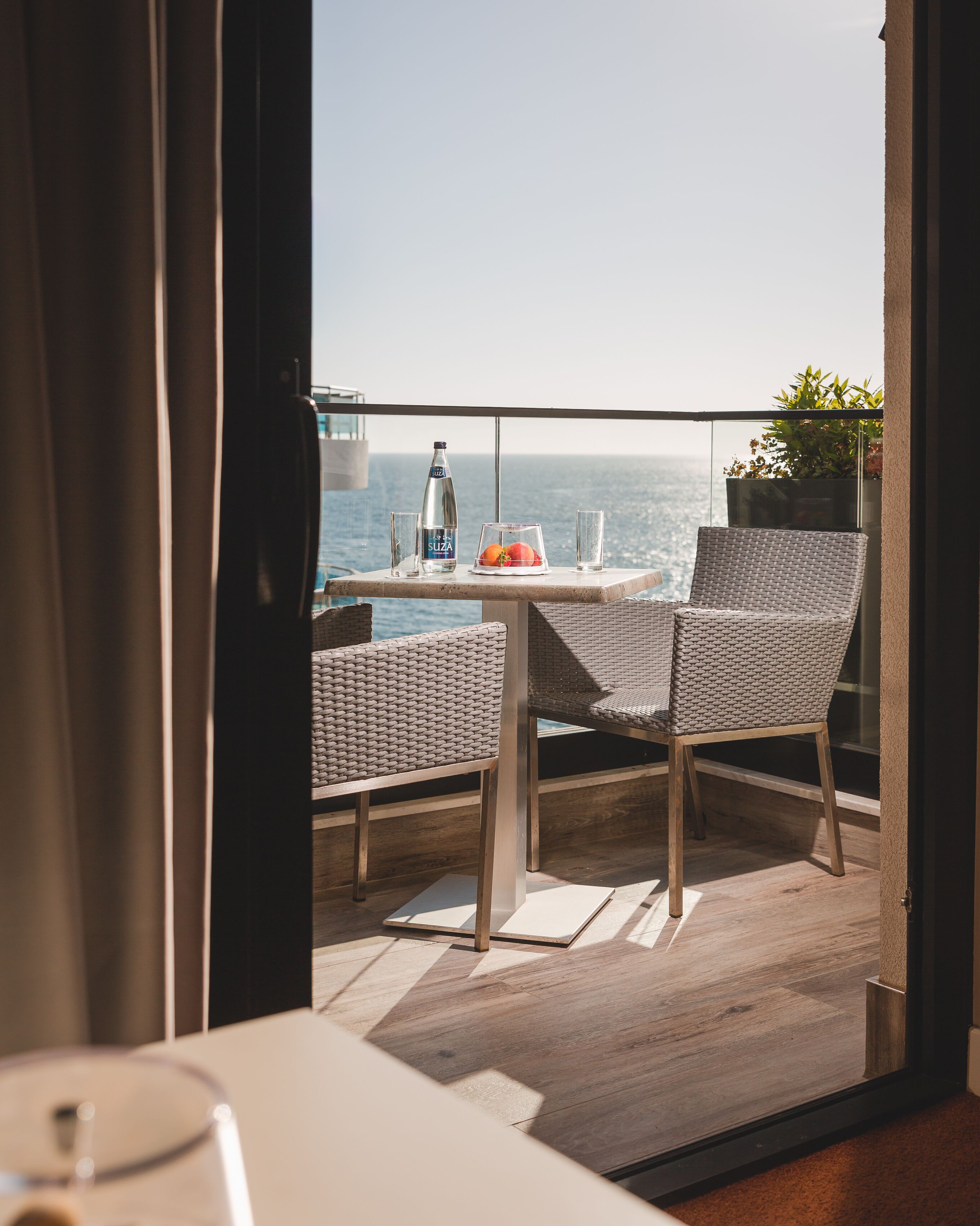 Un balcon avec une table, deux chaises en osier, une bouteille d’eau et un verre.