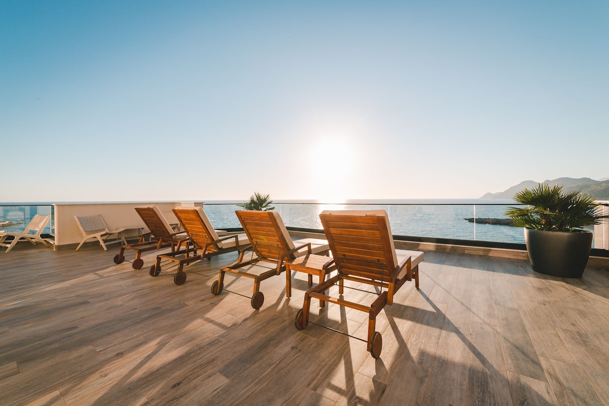 Terraza soleada con sillones de madera y una planta en maceta, con vistas a un cuerpo de agua y montañas distantes.