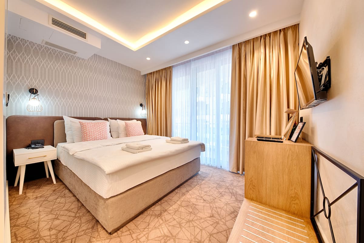 Una habitación de hotel moderna con una cama grande, mesitas de noche, un aparador de madera y un ventanal con cortinas.
