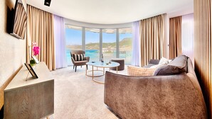 Une chambre d’hôtel moderne avec un plafond incurvé, un canapé, un fauteuil, une petite table et une vue sur la mer.