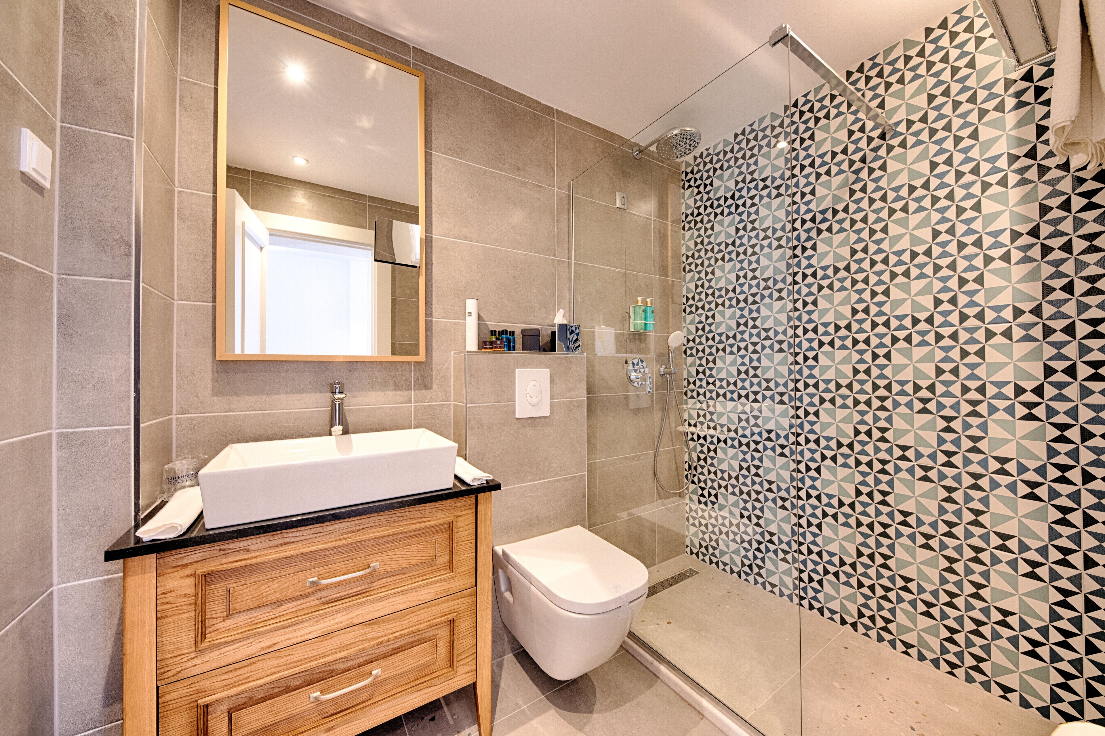 Un baño moderno con un espejo grande, un lavamanos blanco, un mueble de tocador de madera, un inodoro y una ducha con un diseño geométrico de azulejos.